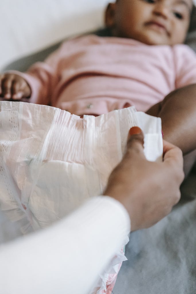 Crop anonymous ethnic mothers holding diaper while dressing calm baby lying on bed at home in daytime